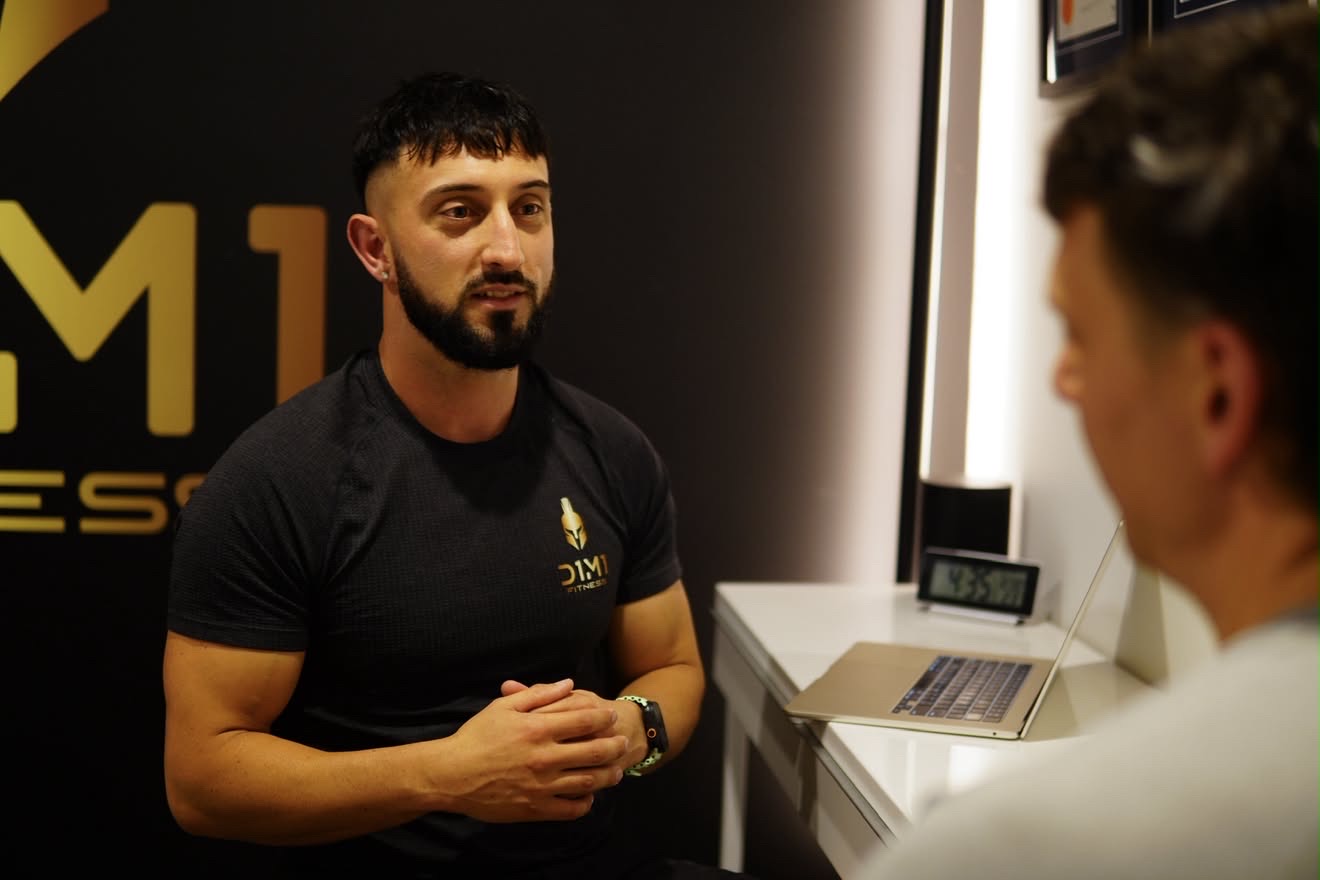 Dimitri Morogiannis consulting with a client at D1M1 Fitness, qualifications framed on the wall